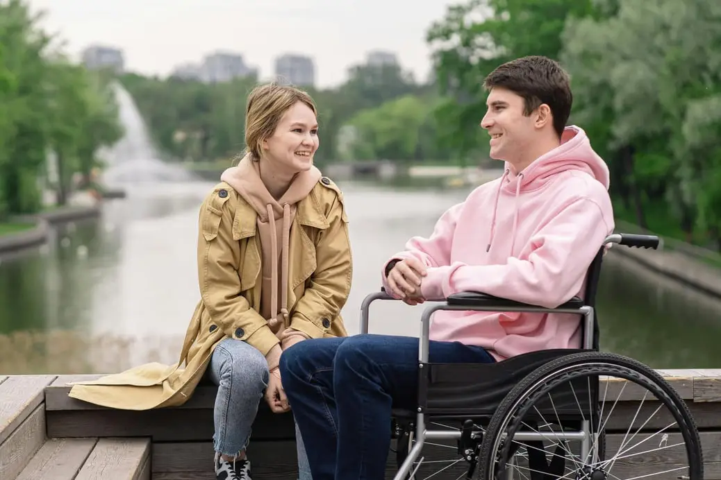 a girl sits on a bench by the river with a boy in a wheelchair in front of her smiling