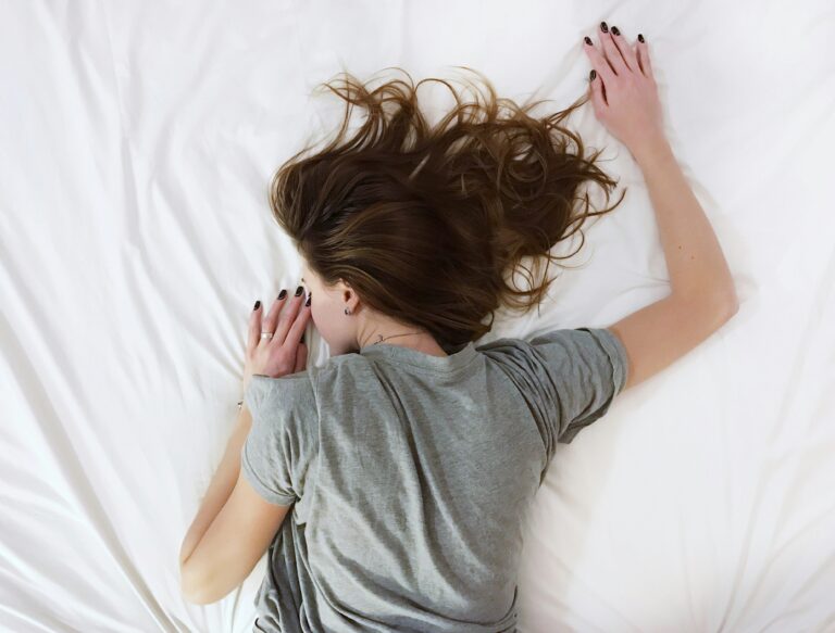 white woman with brown hair and grey shirt lays face down on bed with arms stretched out