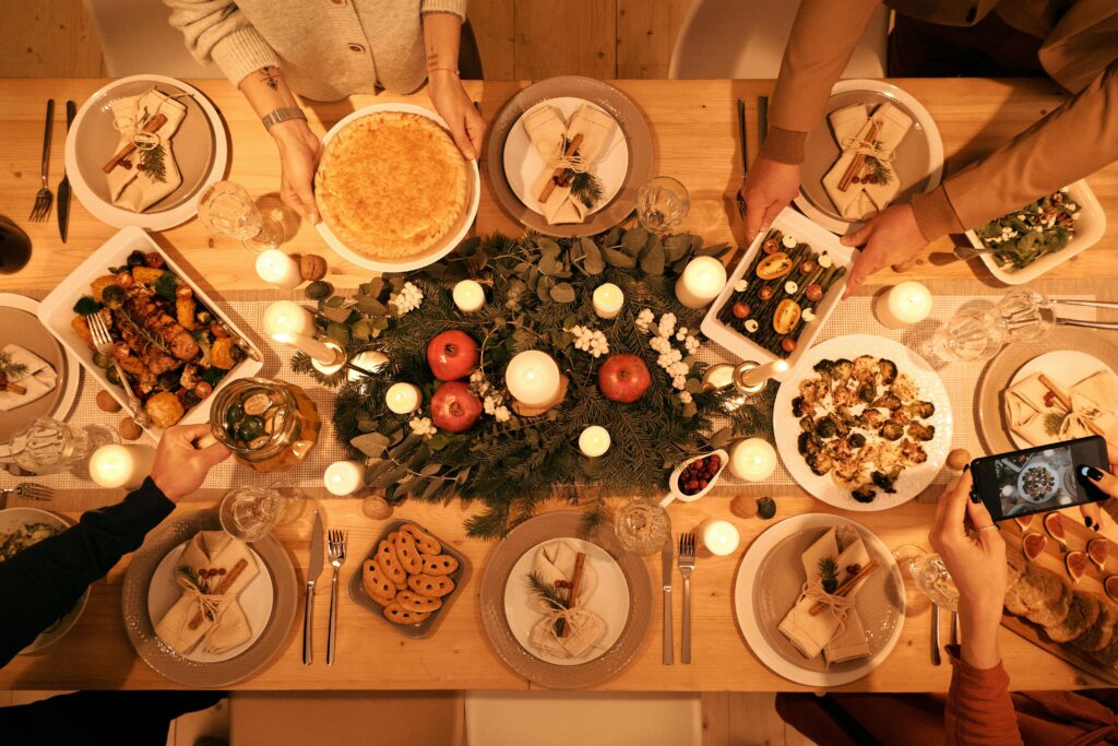 A top down view of a christmas dinner table with multiple dishes and four people with their hands in shot holding dishes, a phone camera and a drink pitcher