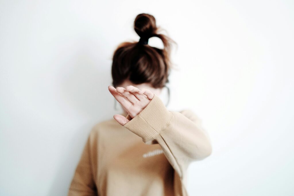 A woman with brown hair in a bun and brown sweater stands against a white background with her hand in front of her face.