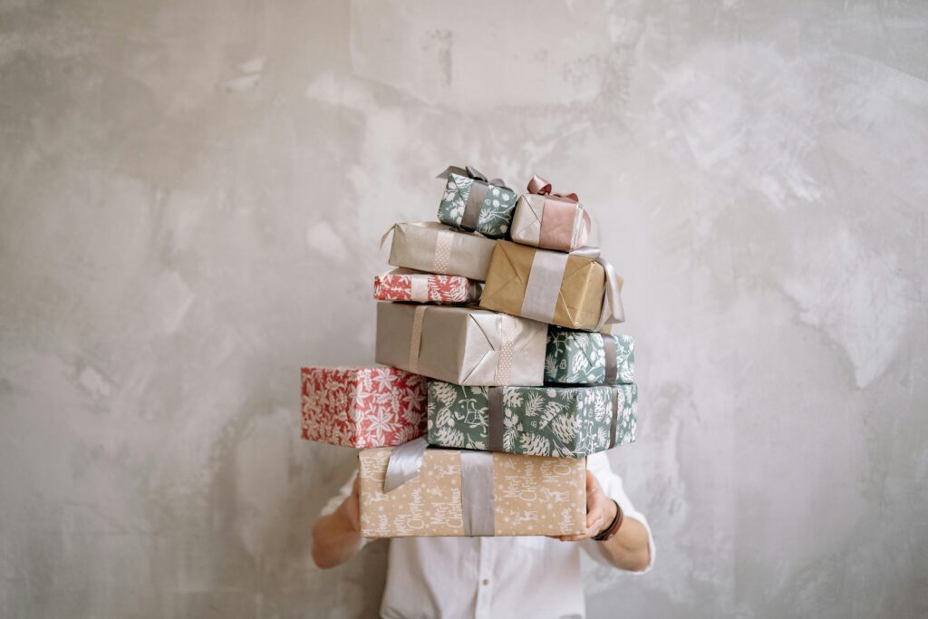 a man holding a stack of 12 gifts in front of their face against a white washed wall