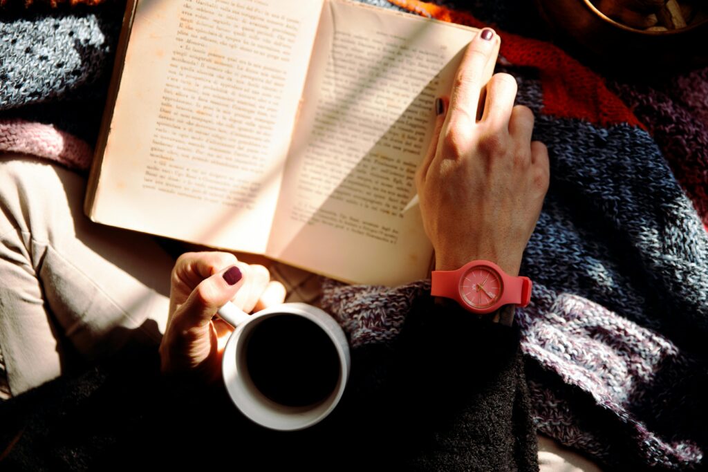 A close up of a womans hands holding a coffee cup and book while wrapped in a blanket.