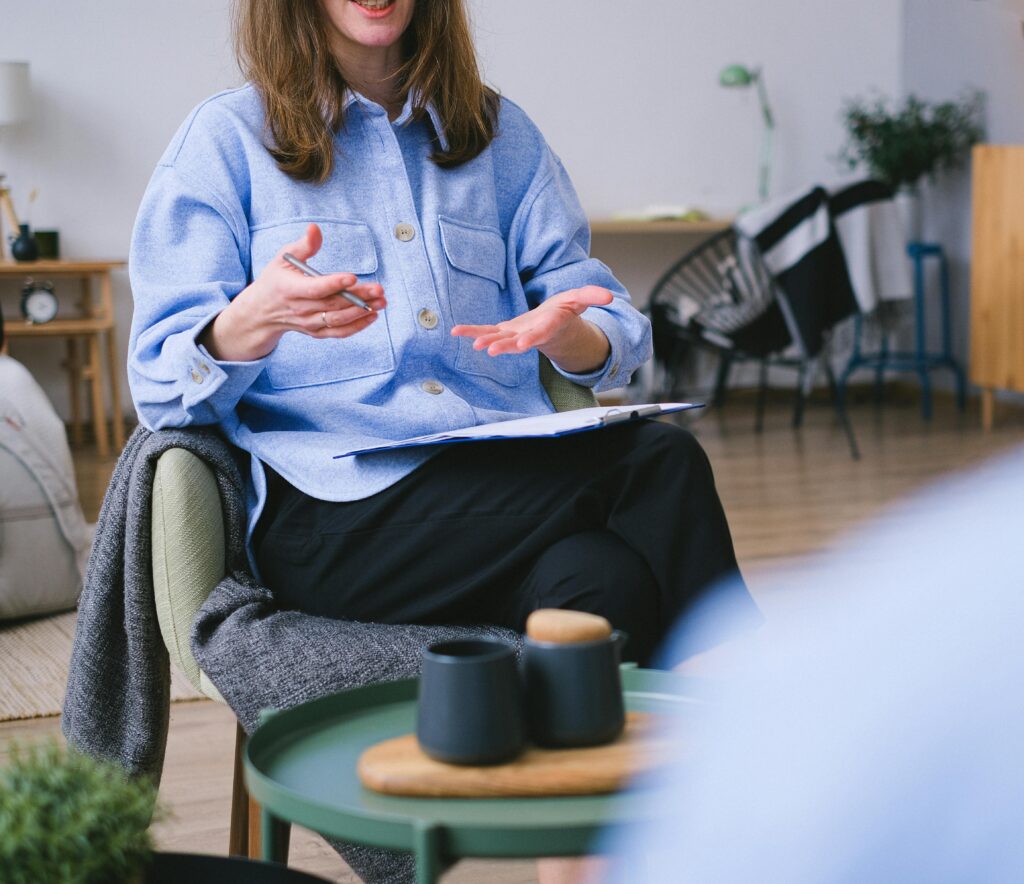 a therapist sits in a chair with a clipboard on her lap, legs cross and she is speaking using her hands.